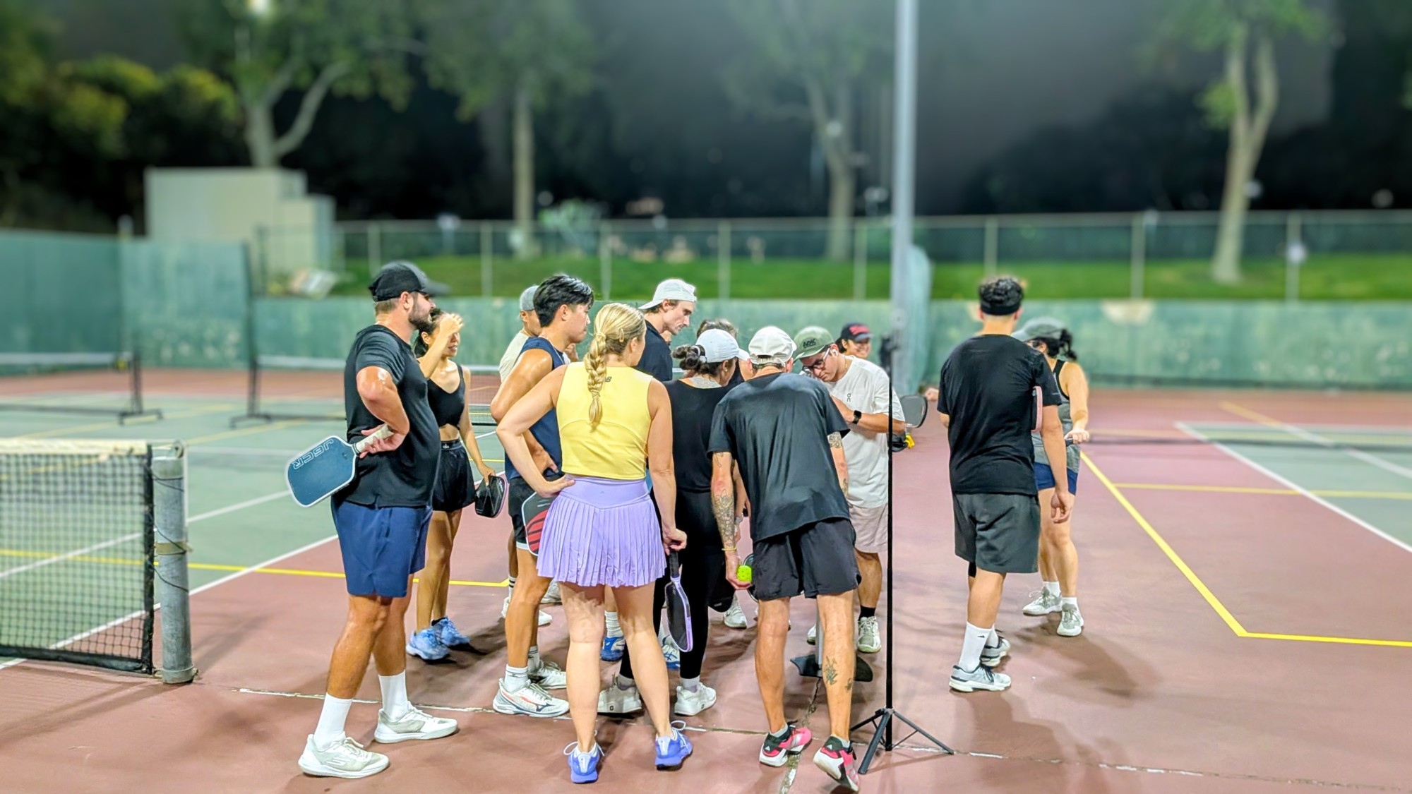 A crowd of players holding paddles gathers around the ShowCourt kiosk between rounds on a lit outdoor court at night